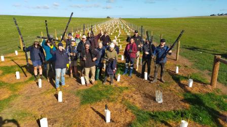 Volunteers from the Phillip Island Landcare group stand on a wide, grassy field, in front of many small plantings protected by plant guards.