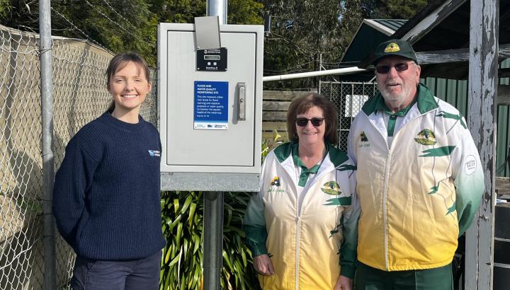 three people standing outdoors next to a digital rain gauge with fencing in background