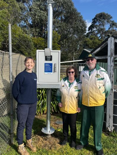 three people standing outdoors next to a digital rain gauge with fencing in background