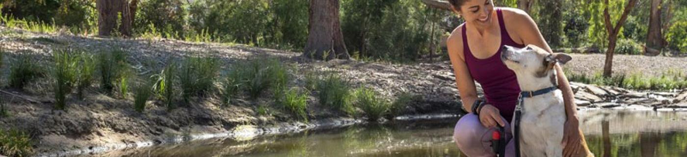 Woman in exercise gear kneels next to her dog on a path across a creek