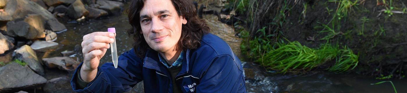 Rhys Coleman crouches by a waterway, holding a test tube.