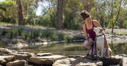 Woman in exercise gear kneels next to her dog on a path across a creek